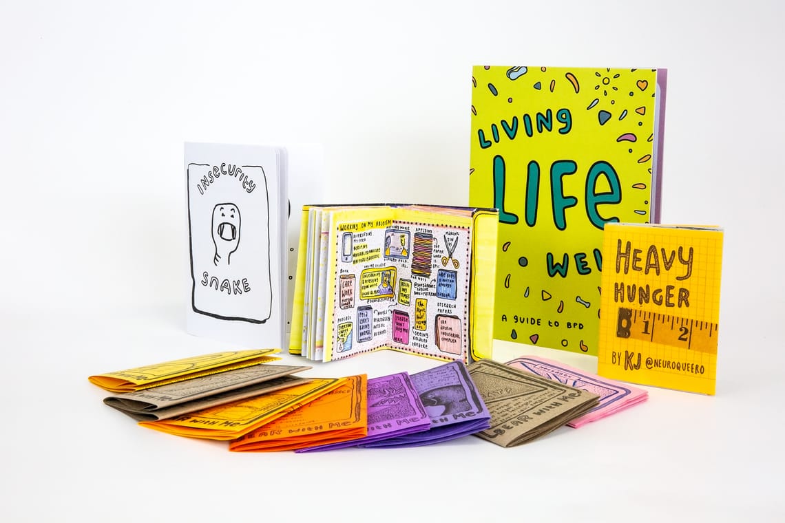 A photo of many zines by KJ Hepworth arranged on a white background. Some are lying down, some are standing up, some are open and some are closed. The zines are hand drawn and colourful.